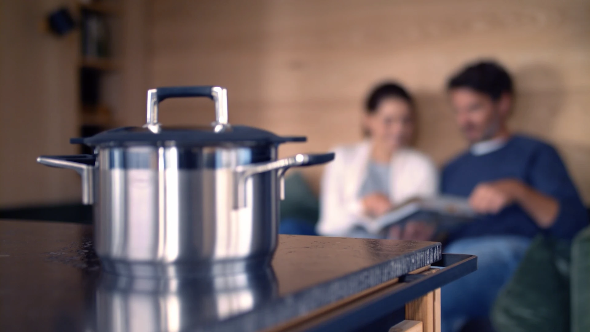 Nahaufnahme eines glänzenden Kochtopfs in moderner Küche, entworfen von Bürobewegt, im Hintergrund unscharfe Personen.