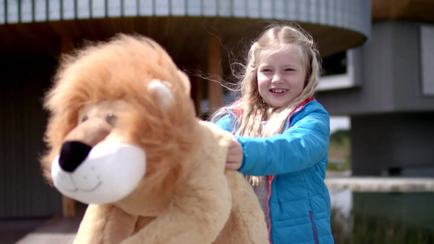 Smiling girl in blue jacket holds a large plush lion outdoors, showcasing Bürobewegt's creative visual marketing.