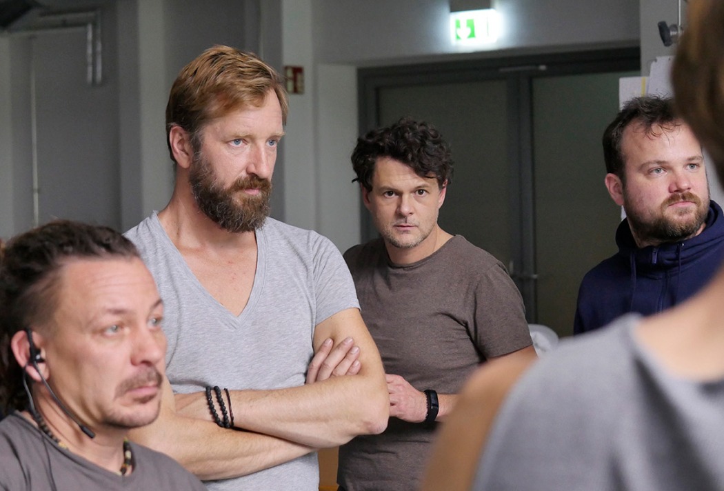 Vier Männer in einem Raum, die konzentriert schauen. Eine Tür und Notausgangsschild sind im Hintergrund sichtbar.