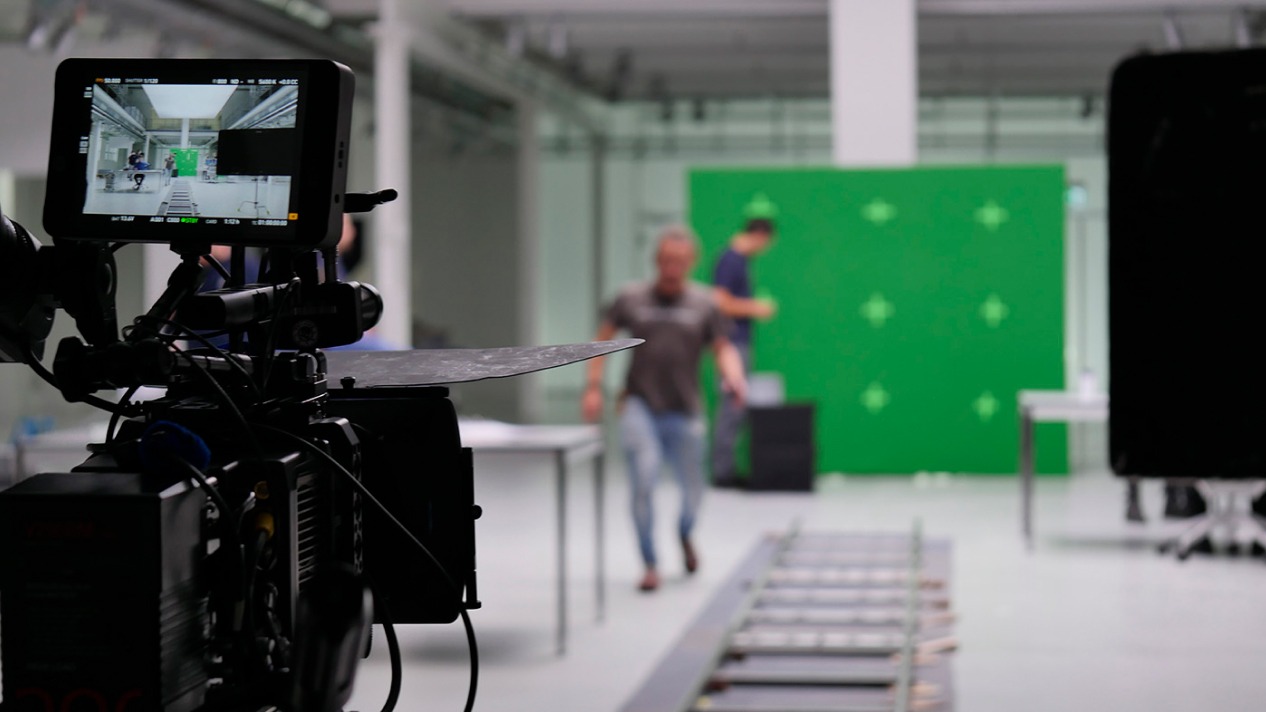 Filmset mit Kamera und Greenscreen im Hintergrund, Personen in Aktion, Fokus auf Videoproduktion.