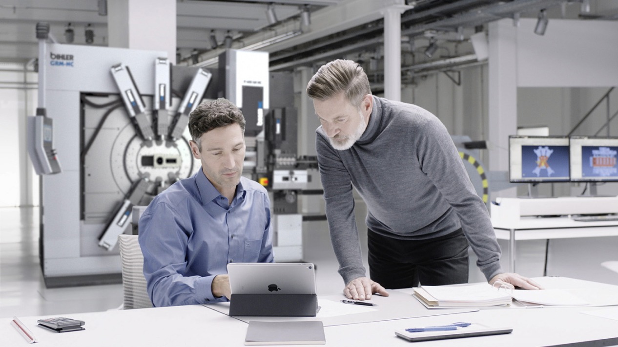 Zwei Männer arbeiten in einem modernen Büro mit Tablet und Maschinen im Hintergrund. Technologie und Zusammenarbeit.