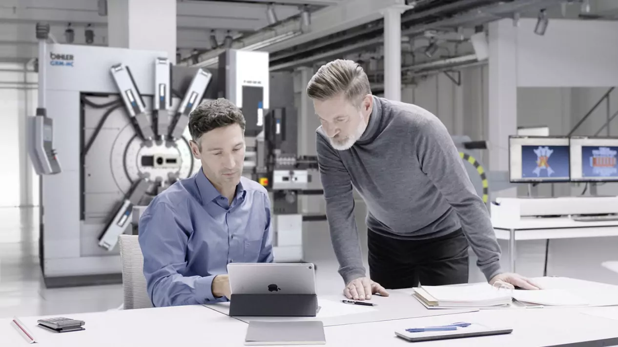 Zwei Männer arbeiten in einem modernen Büro mit Tablet und Maschinen im Hintergrund. Technologie und Zusammenarbeit.