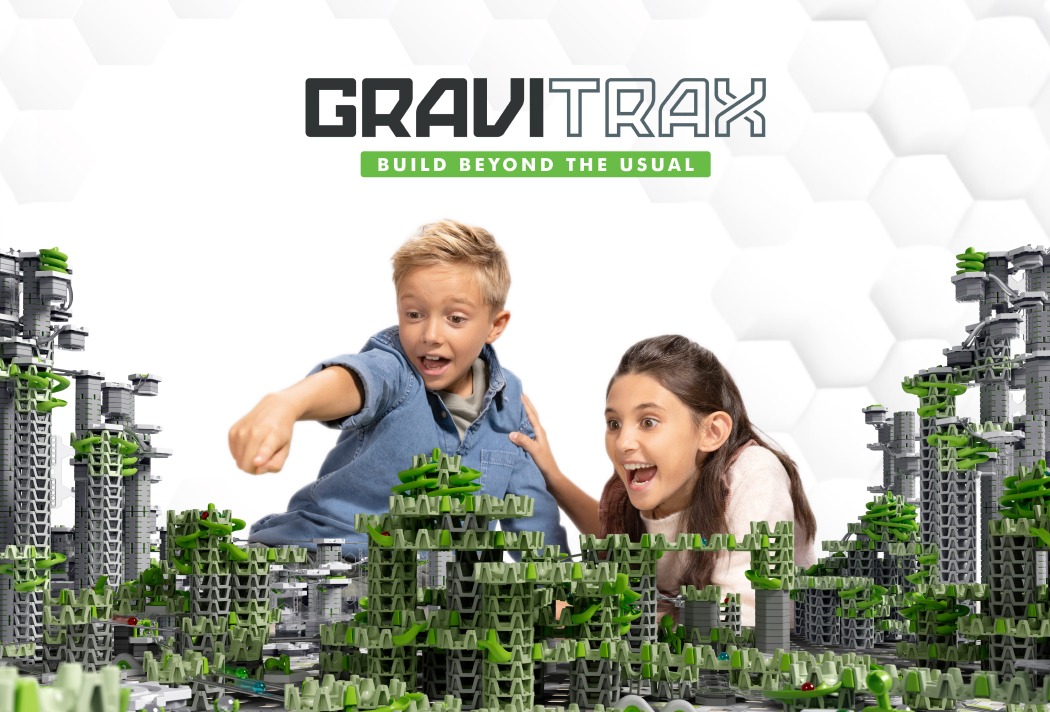 Zwei Kinder spielen begeistert mit einer GraviTrax-Kugelbahn mit grünen und grauen Bauteilen vor einem weißen Hintergrund.