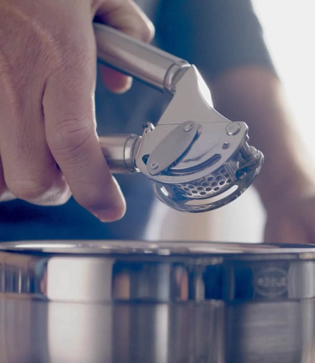 Hand benutzt Knoblauchpresse über Kochtopf in der Küche. Nahaufnahme der Edelstahl-Küchenwerkzeuge.