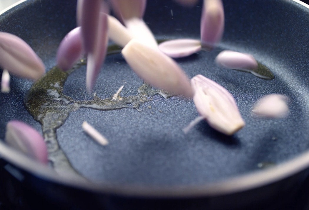 Zwiebelstücke fallen in eine Pfanne mit Öl zum Anbraten, Nahaufnahme der Kochszene in Bewegung.