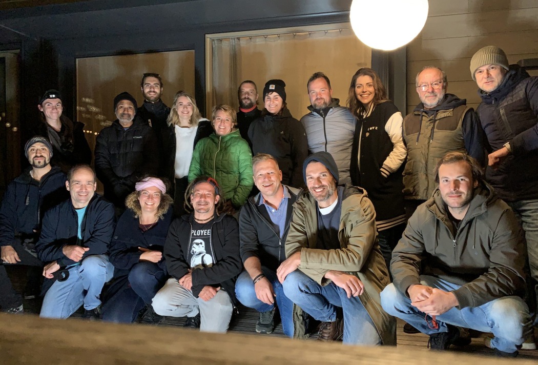 Gruppe von 18 Menschen lächelt in die Kamera, gekleidet für kühleres Wetter, versammelt vor einem Gebäude.