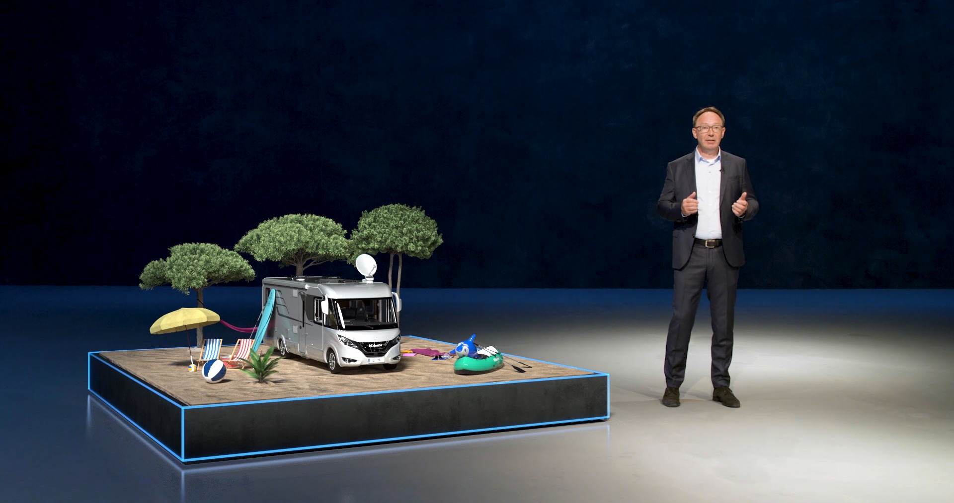 Präsentation eines Wohnmobils auf Bühne mit Urlaubsdekoration, Sprecher im Anzug erklärt Funktionen.
